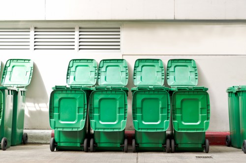 A row of six large green plastic wheelie bins with black wheels and closed lids, positioned on a paved surface against a white building wall with horizontal ventilation slots above. The bins are aligned in a neat, single file with some slightly behind others, and the surface appears clean and well-maintained. In the background, the building wall extends to the right with more green bins partially visible outside the frame. The setting suggests an outdoor area typical for waste collection and rubbish removal services, possibly in an urban or commercial environment in Barbican or nearby London locations. The natural daylight creates soft shadows on the ground, emphasizing the texture of the bins' lids and bodies, highlighting their robust plastic construction, suitable for waste storage or disposal purposes associated with commercial waste removal activities by companies like Commercial Waste Removal Barbican.