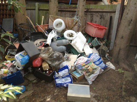 A large pile of mixed household and garden waste situated outdoors on bare soil, including white plastic pipes, a red plastic storage bin, black plastic containers, and various cardboard boxes. The waste also comprises crumpled plastic bags, paper packaging, and miscellaneous debris scattered unevenly across the area, with some items leaning against a weathered wooden fence and trees in the background. The overall scene reflects an accumulation of rubbish ready for removal, consistent with commercial waste collection services such as those provided by Commercial Waste Removal Barbican, serving local business and property owners in the Barbican area. The environment appears to be a backyard or side courtyard with natural light, and the waste materials vary in texture from smooth plastic to coarse cardboard and paper, highlighting the need for professional clearance to maintain cleanliness and compliance with waste disposal regulations in the city center.