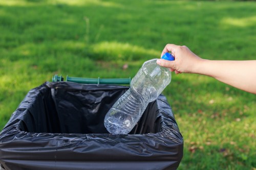 A person's hand is seen dropping a clear plastic water bottle into a black bin liner placed inside a large, open-sided rubbish bin outdoors. The bin is situated on a paved surface with a grassy area and trees visible in the background, indicating a park or garden environment. The hand holds the water bottle by the neck, with the bottle appearing partially crushed and transparent, revealing the water inside. The rubbish bin's metal exterior and its open top are visible, with the bin liner lining the inside, appearing loose and slightly crumpled around the edges. The scene is brightly lit with natural daylight, emphasizing the outdoor setting. The image is relevant to waste management services provided by Commercial Waste Removal Barbican, emphasizing responsible rubbish disposal practices in a residential or commercial outdoor space near Barbican.