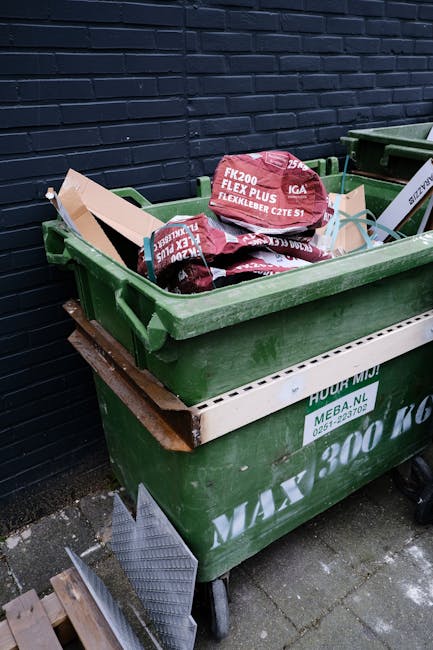 A row of upright wheelie bins made of plastic, positioned outdoors along a pavement beside a brick wall. The bins are in various colours, with green and blue in the foreground, and red and dark green 